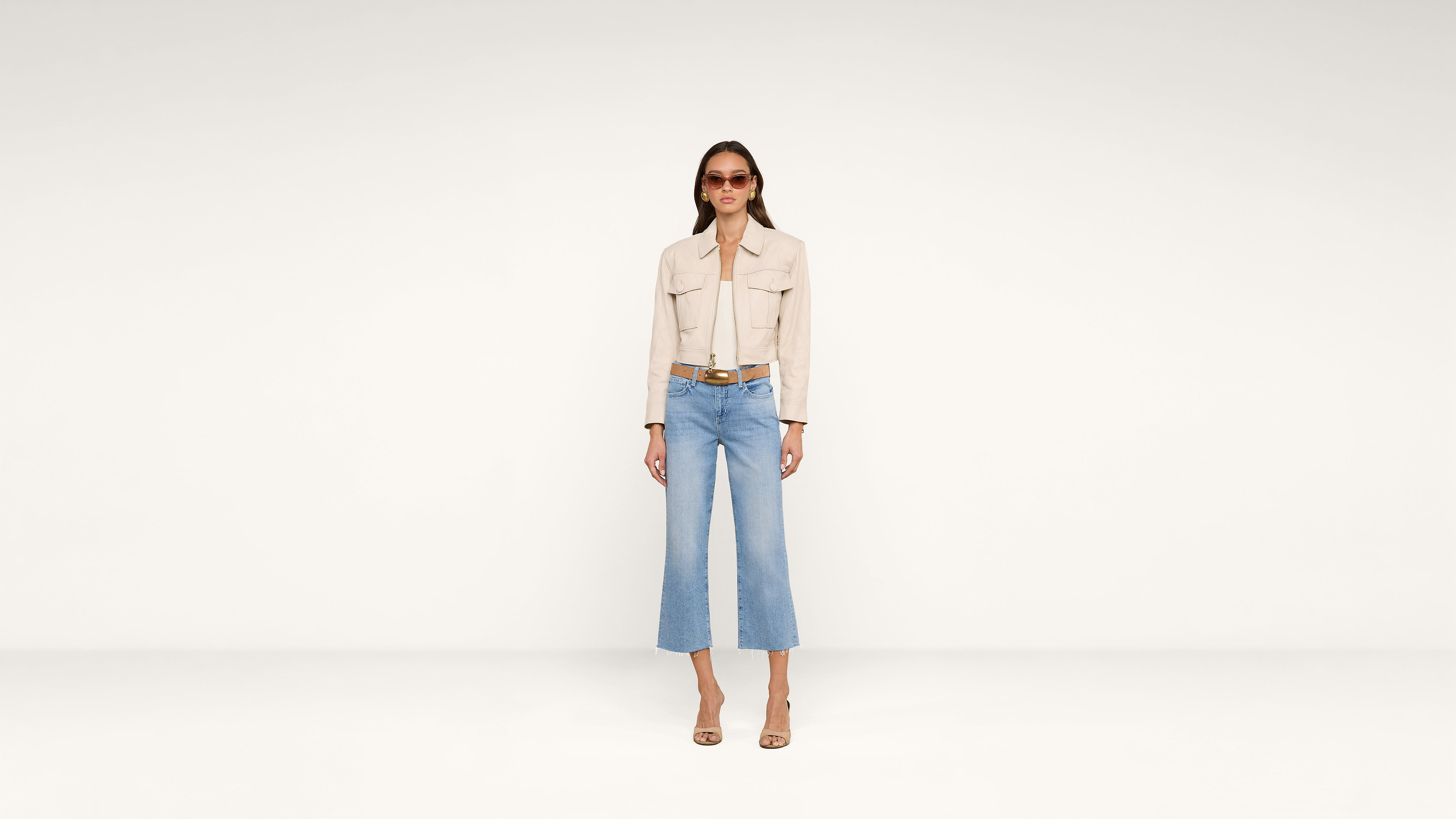 A woman wearing sunglasses, a beige jacket, light blue cropped jeans, and heeled sandals stands against a plain white background.