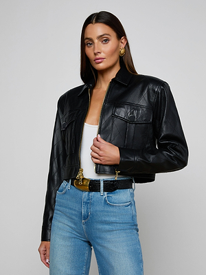 A woman wears the Micah Oversized Leather Jacket in black over a white top and light blue jeans, styled with gold earrings and a black belt, standing against a plain light gray background.