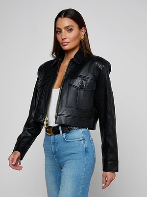 A woman with long brown hair wears the Micah Oversized Leather Jacket over a white top and blue jeans, paired with gold hoop earrings, standing against a plain light background.