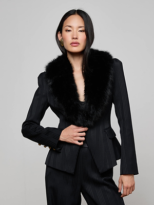 A woman with long dark hair wears the L'AGENCE Kaisley Faux Fur Collar Blazer, a fitted black pinstripe suit with a wide faux fur collar, paired with gold hoop earrings, posing confidently against a light gray background.