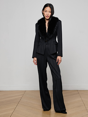 A woman with straight dark hair stands on a parquet floor, wearing the L'AGENCE Kaisley Faux Fur Collar Blazer—a fitted, pinstripe black suit jacket featuring a stylish faux fur collar.