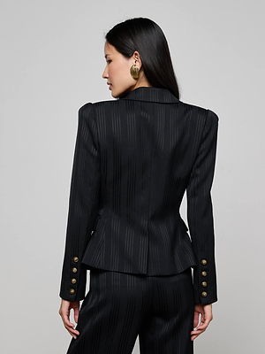 A woman with long dark hair and large round earrings faces away from the camera, wearing the L'AGENCE Kaisley Faux Fur Collar Blazer. The black pinstripe blazer features gold cuff buttons. The background is plain and light-colored.