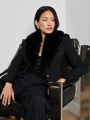 A woman with long dark hair, wearing the L'AGENCE Kaisley Faux Fur Collar Blazer and gold earrings, sits confidently in a modern black leather and metal chair, looking to the side.