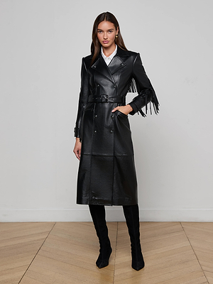 Indoors against a plain light wall, a woman wears the Celina Fringed Leather Trench—a long black lambskin coat with fringed sleeve trim—over a white collared shirt and black knee-high boots, accessorized with minimal jewelry.