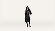 A woman wearing a long black leather trench coat and black boots stands against a plain white background.