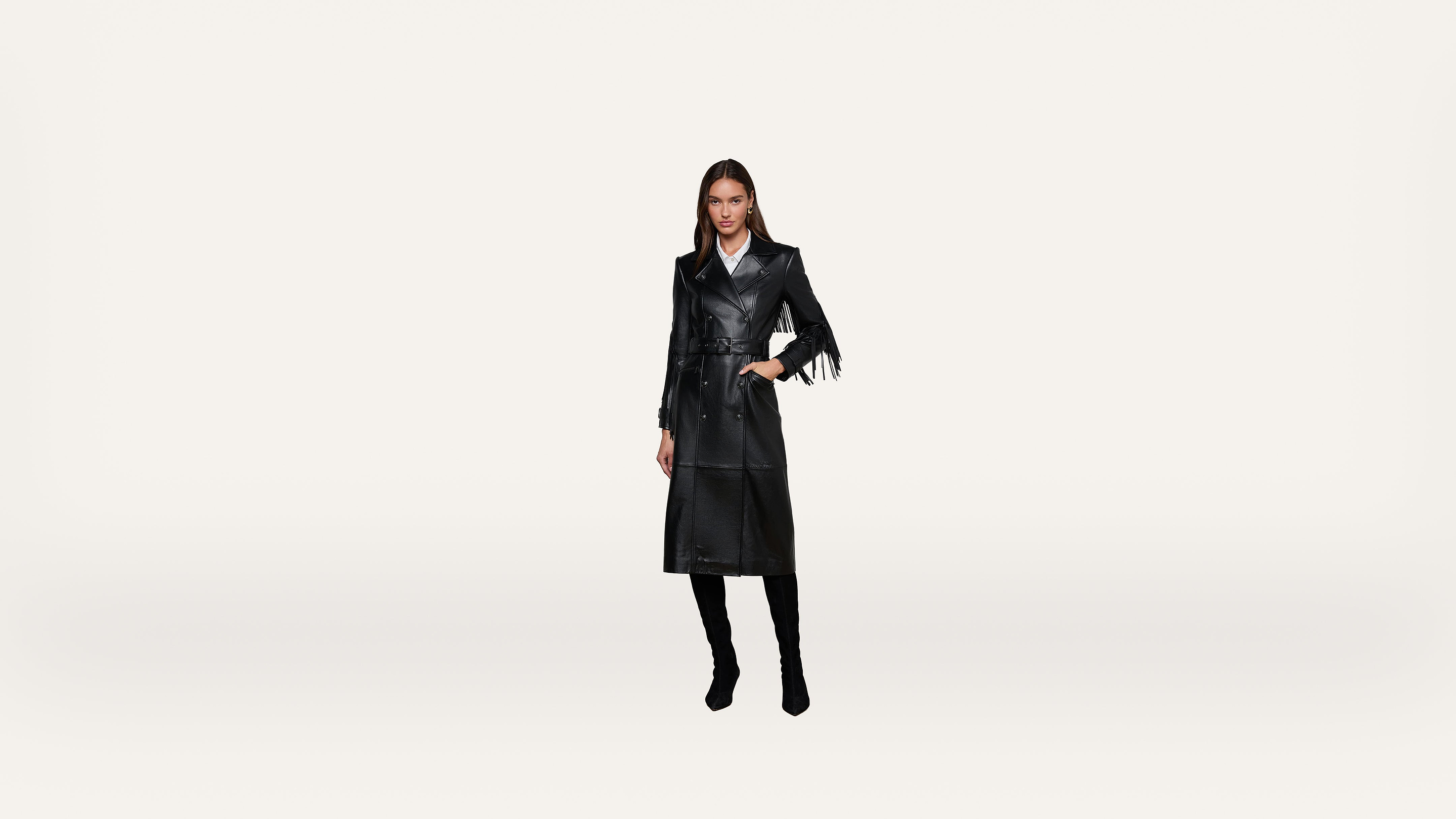 A woman wearing a long black leather trench coat and black boots stands against a plain white background.