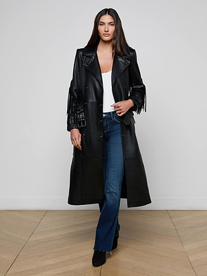 A woman stands indoors against a plain light wall wearing the Celina Fringed Leather Trench—a long black lambskin coat with fringed sleeve trim—paired with a white top, blue jeans, and black heeled boots. She has straight dark hair.
