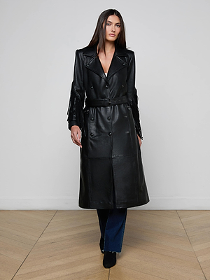 A woman with long straight hair and a neutral expression wears the Celina Fringed Leather Trench—a long black lambskin coat with a belted waist—over dark blue jeans and black boots, standing indoors against a plain light-colored wall.