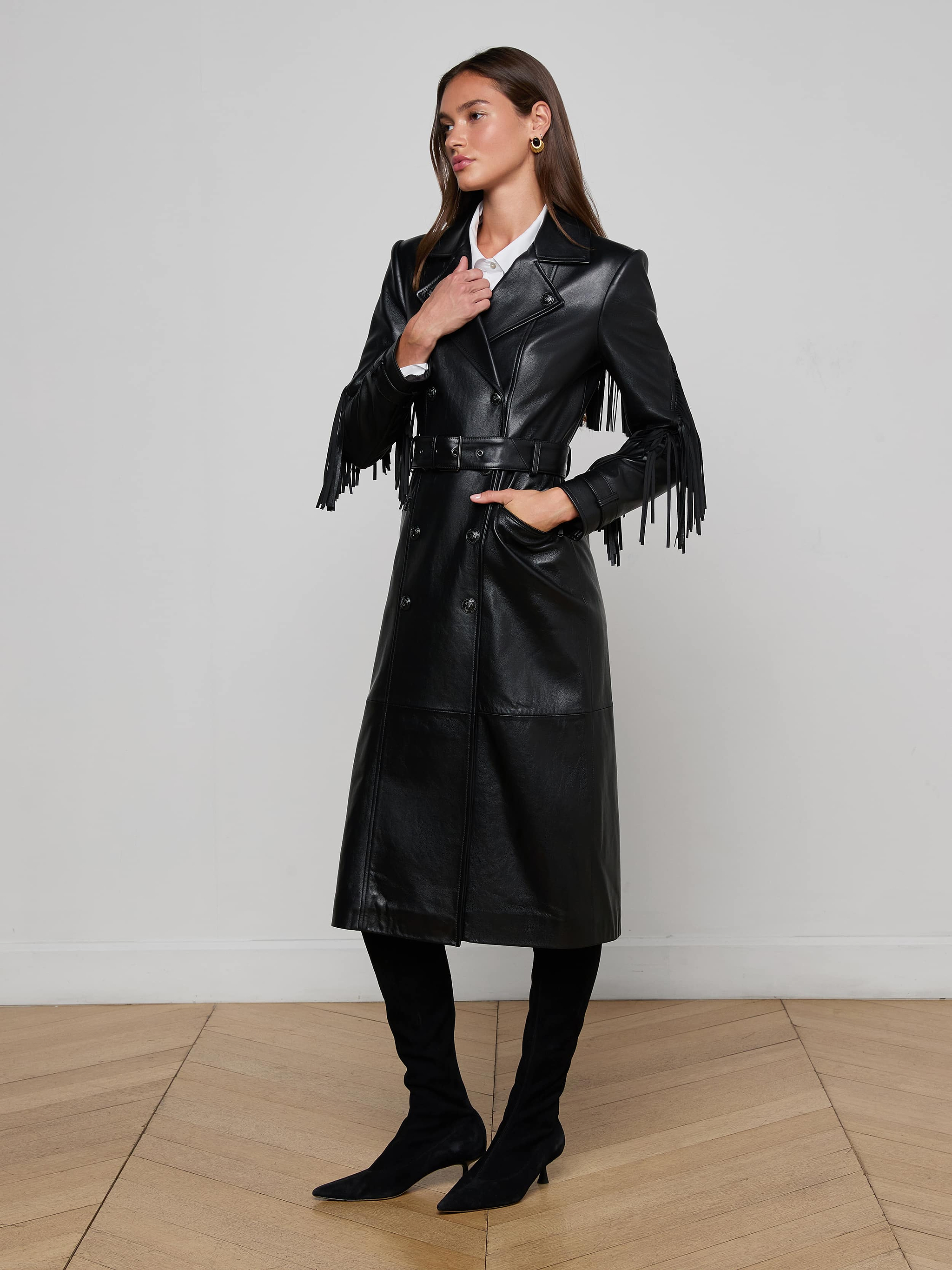Indoors on a wooden floor, a woman models the Celina Fringed Leather Trench—a long black leather coat with fringed trim—paired with a white collared shirt, black knee-high boots, and gold earrings. One hand rests on her neck, the other in her pocket.