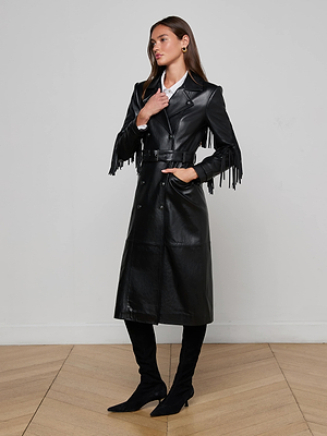 Indoors on a wooden floor, a woman models the Celina Fringed Leather Trench—a long black leather coat with fringed trim—paired with a white collared shirt, black knee-high boots, and gold earrings. One hand rests on her neck, the other in her pocket.