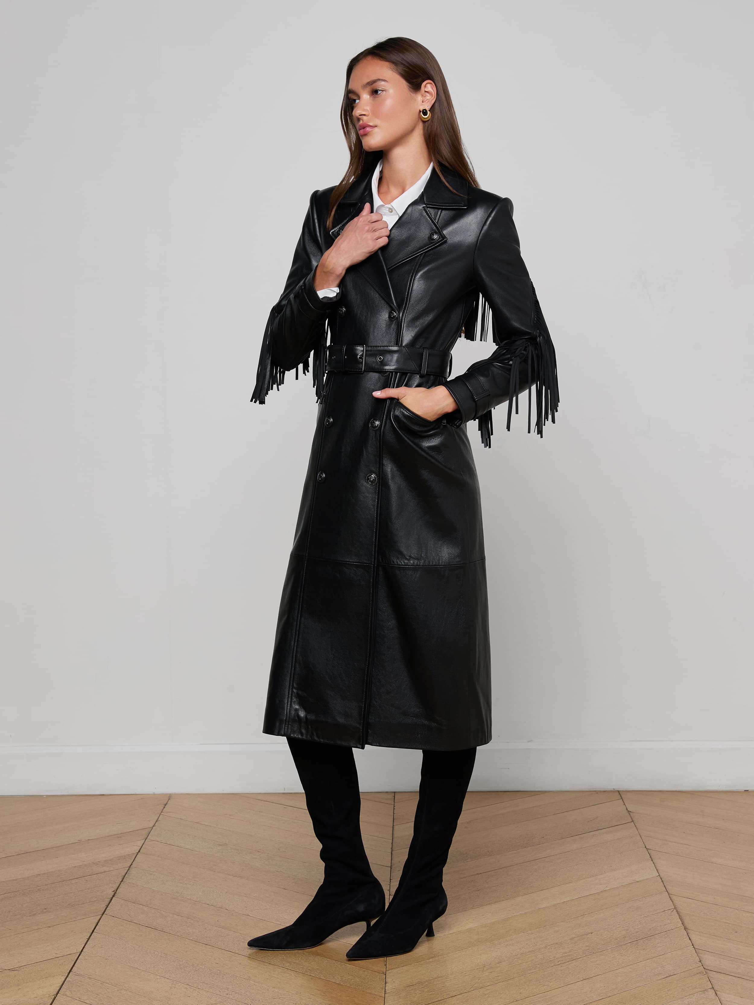 Indoors on a wooden floor, a woman models the Celina Fringed Leather Trench—a long black leather coat with fringed trim—paired with a white collared shirt, black knee-high boots, and gold earrings. One hand rests on her neck, the other in her pocket.
