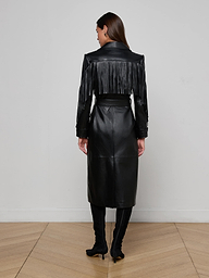 A woman faces away, wearing the Celina Fringed Leather Trench—black leather with fringe across the upper back and arms—paired with black heeled boots in a minimalist room featuring light wood floors and a plain white wall.