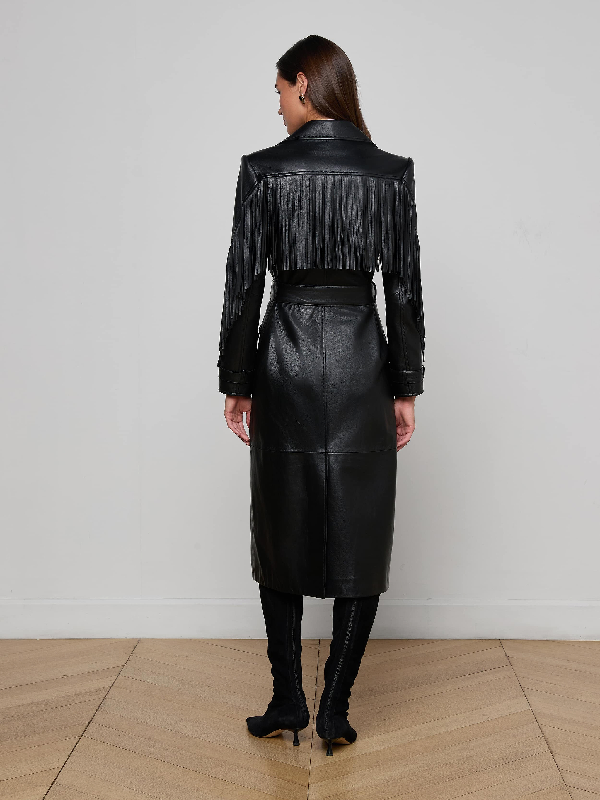 A woman faces away, wearing the Celina Fringed Leather Trench—black leather with fringe across the upper back and arms—paired with black heeled boots in a minimalist room featuring light wood floors and a plain white wall.