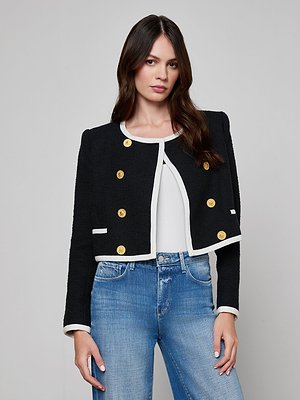 A woman wears the Mazikeen Tweed Jacket—black, cropped, with gold buttons and white trim—over a white top and blue jeans, standing against a plain light gray background.