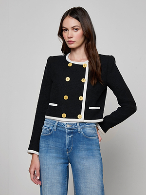 A woman with long dark hair models the Mazikeen Tweed Jacket—black, cropped, with gold buttons and white trim—paired with blue jeans. She stands against a light gray background, one hand on her hip.