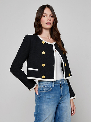 A woman wears the Mazikeen Tweed Jacket in black with white trim and gold buttons over a white top and blue jeans, standing against a light gray background.