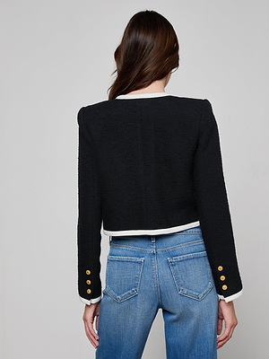 A woman with long brown hair, seen from behind, wears the Mazikeen Tweed Jacket—black with gold sleeve buttons and white trim—paired with blue jeans against a plain light gray background.