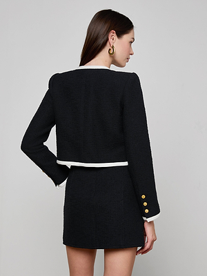 A woman with long brown hair models the Mazikeen Tweed Jacket, featuring white trim and gold sleeve buttons, paired with a matching black skirt. She stands with her back to the camera against a plain light background.