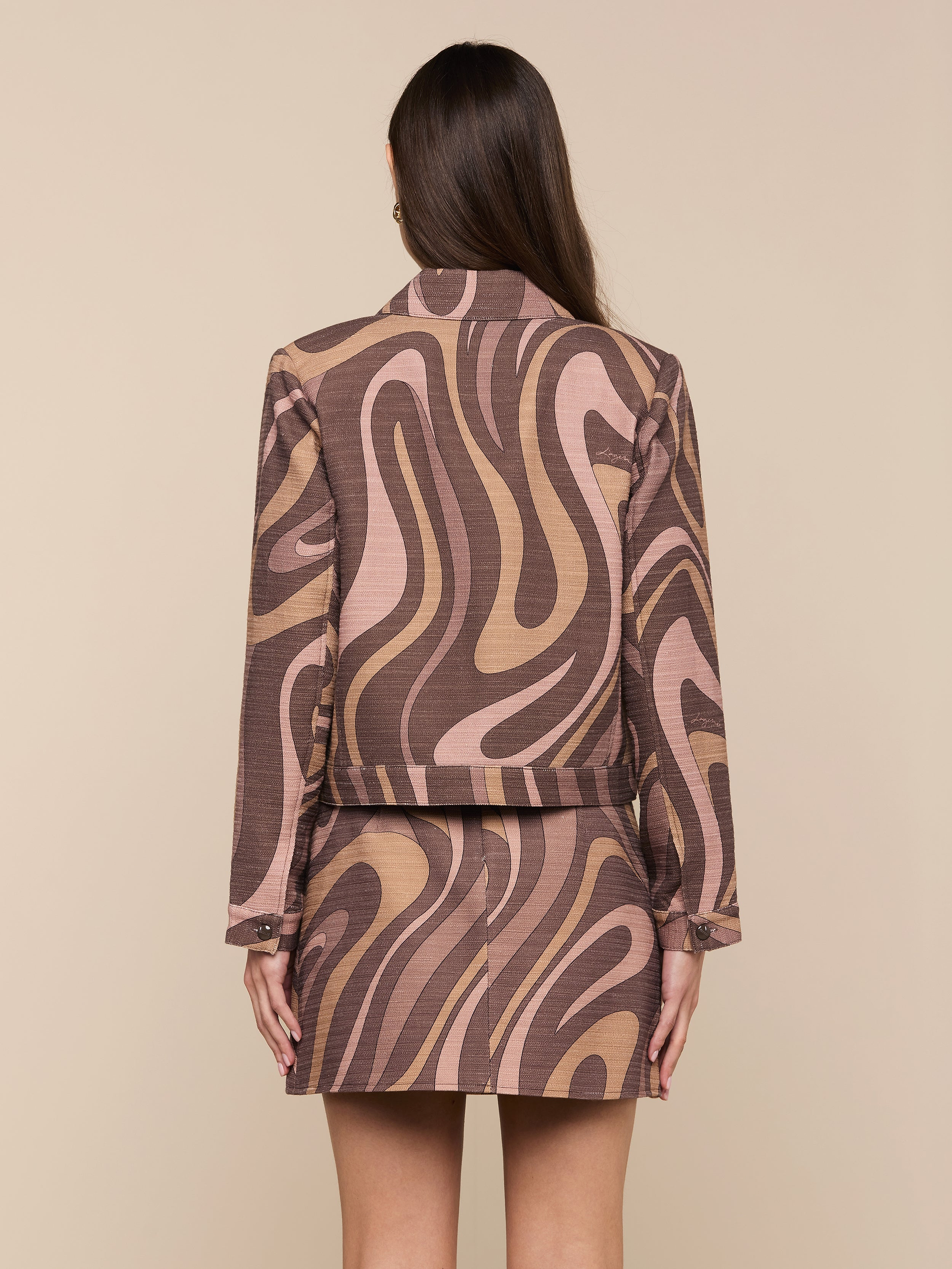 A woman with long brown hair faces away, wearing the Nyx Cropped Jacket featuring an abstract swirl print and a matching skirt. Her arms are relaxed by her sides as she stands against a plain light background.