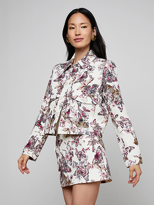 A woman with long black hair wears the Nyx Cropped Jacket—a lightweight printed cotton piece featuring a vibrant butterfly jungle print—paired with a matching skirt, standing against a plain light gray background.