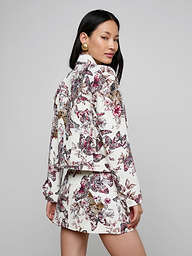 A woman with long dark hair and gold earrings poses in the Nyx Cropped Jacket featuring a matching butterfly jungle print, with her body turned to the side and looking over her shoulder against a neutral background.