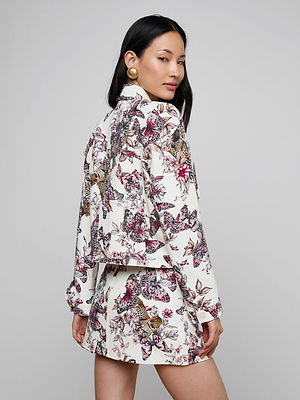 A woman with long dark hair and gold earrings poses in the Nyx Cropped Jacket featuring a matching butterfly jungle print, with her body turned to the side and looking over her shoulder against a neutral background.
