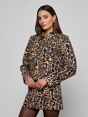 A woman with long straight hair and gold earrings stands against a plain background, wearing the Nyx Cropped Jacket in relaxed fit leopard print with large gold buttons. She has a neutral expression and her hands gently clasped in front.
