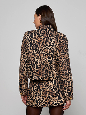 A woman with long brown hair is seen from the back, wearing the Nyx Cropped Jacket in a relaxed fit leopard print with a matching skirt and black tights. The background is plain and light-colored.