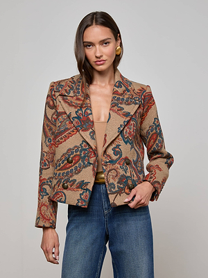 A woman wearing the L'AGENCE Kelsey Peacoat with intricate red, blue, and brown designs, paired with blue jeans and gold earrings, stands against a plain light background, radiating vintage charm.