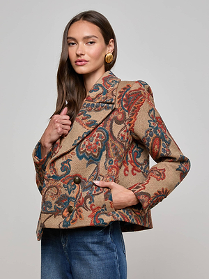 A woman models the Kelsey Peacoat by L'AGENCE, a brown paisley jacquard blazer with blue and red accents, paired with blue jeans and gold earrings, set against a plain gray backdrop.