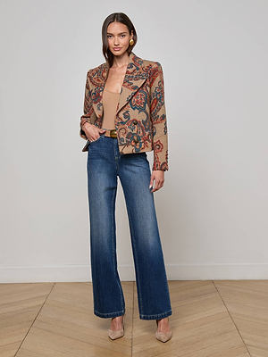 A woman with long brown hair, smiling slightly, poses with one hand in her pocket on a wood floor. She wears the L'AGENCE Kelsey Peacoat, a tan top, blue high-waisted wide-leg jeans, and beige heels.