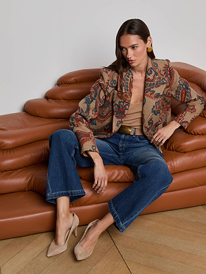 A woman sits on a brown leather sofa, wearing blue jeans, a tan top, tan heels, gold earrings, and the Kelsey Peacoat by L'AGENCE. She rests one arm on the sofa and crosses her legs at the ankle.