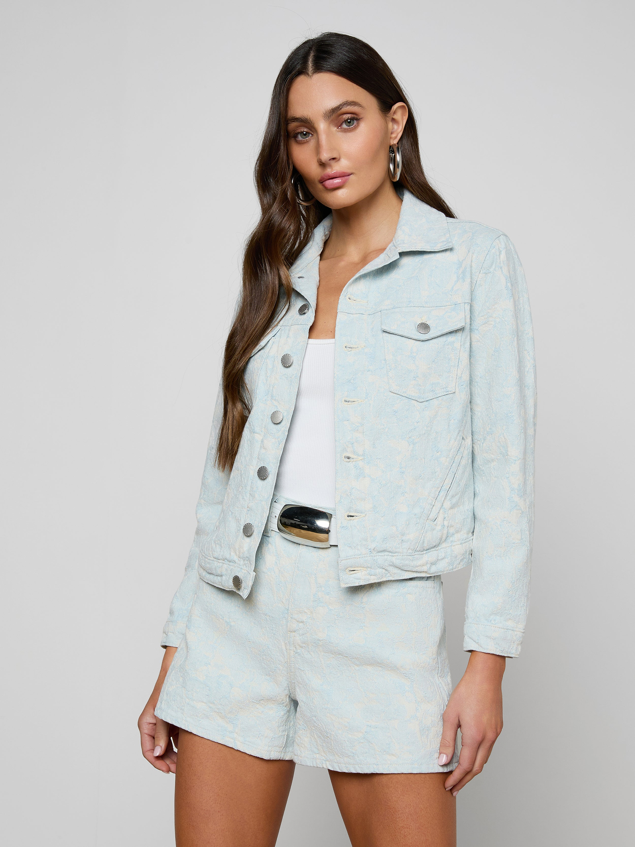 The woman wears the Maven Jacket—a structured fit, light blue denim piece with three-quarter sleeves—paired with matching shorts over a white top. Her long brown hair flows down as she stands against a plain, light background.