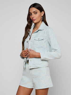 A woman with long brown hair wears the light blue cotton jacquard Maven Jacket and matching shorts, standing against a plain light gray background. She faces slightly sideways, holding the jacket’s front with her hands.