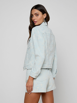 A woman with long brown hair wears the Maven Jacket and matching shorts in light blue cotton jacquard, standing sideways and looking over her shoulder toward the camera against a plain light gray background.