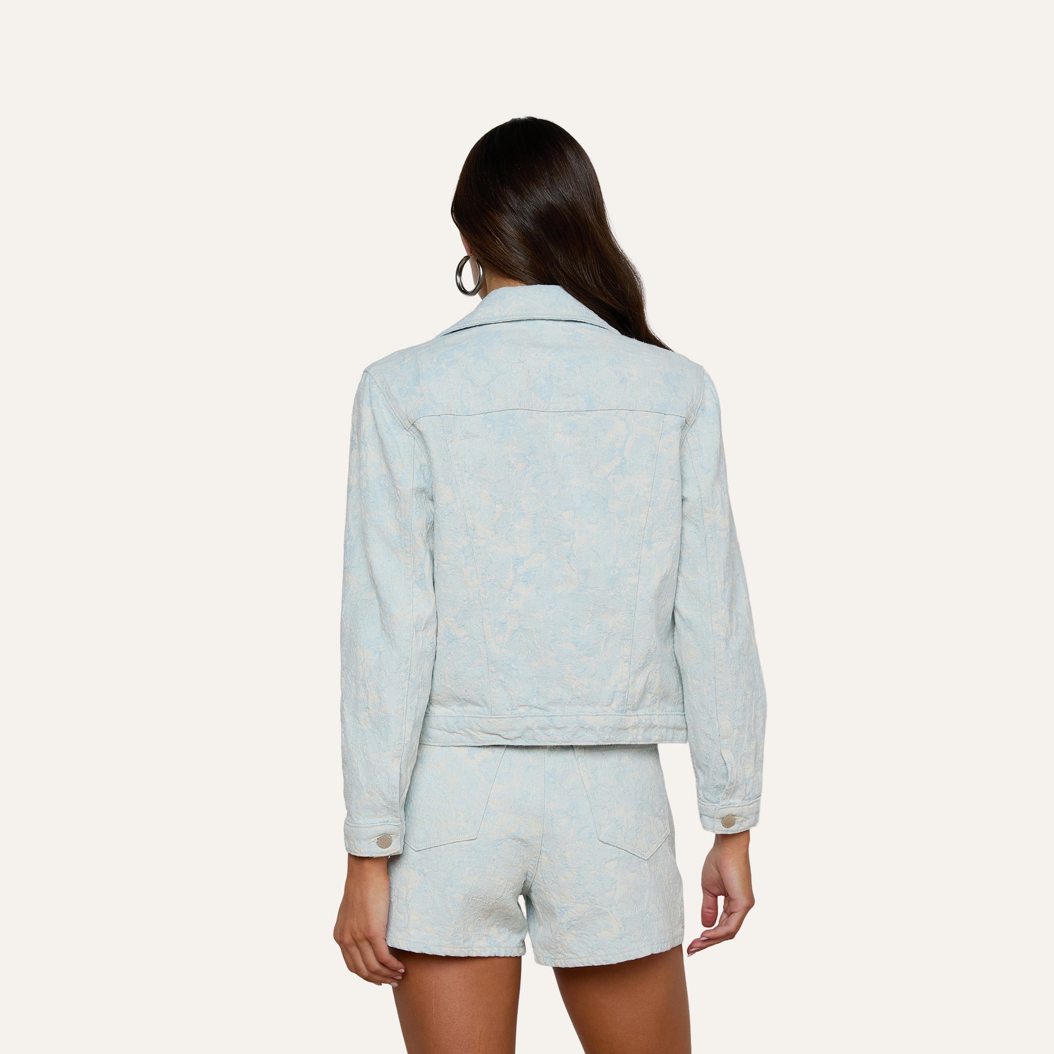 A woman with long brown hair is shown from behind, wearing the Maven Jacket in light blue with matching shorts. The background is plain and off-white.