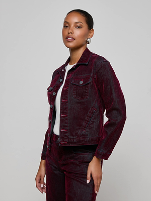 A woman models the L'AGENCE Maven Flocked Jean Jacket—a dark red, velvet-finished piece—paired with matching pants. She stands against a light gray background, hair pulled back, confidently facing the camera.