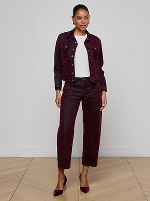 A woman poses indoors against a light wall, wearing the L'AGENCE Maven Flocked Jean Jacket in burgundy with matching pants, a white top, and burgundy heels, standing on a wooden floor.