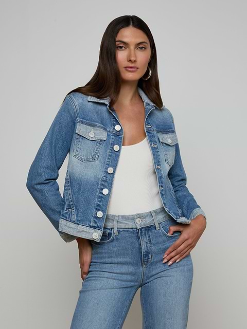 A woman stands against a plain light background with one hand on her hip, wearing the Maven Jean Jacket—a light-wash, three-quarter sleeve jean jacket—over a white top and blue jeans.
