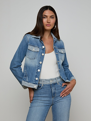 A woman with long brown hair wears the Maven Jean Jacket over a white top and matching blue jeans, standing against a plain light background with one hand on her hip and a neutral expression.