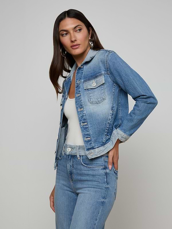 A woman with long dark hair wears the Maven Jean Jacket over a white top and matching blue jeans, standing against a plain light background, with one hand on her hip and looking slightly to the side.