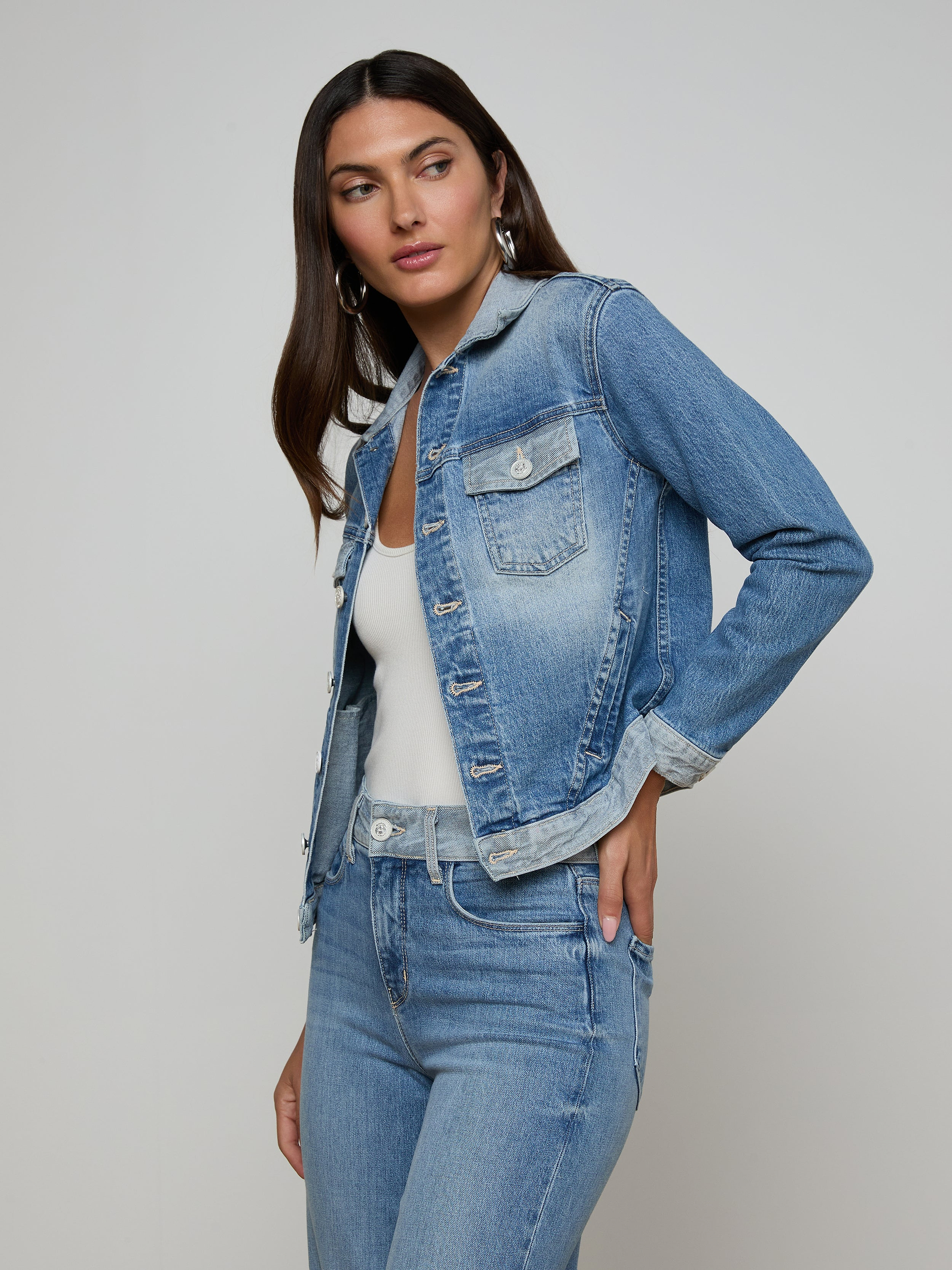A woman with long dark hair wears the Maven Jean Jacket over a white top and matching blue jeans, standing against a plain light background, with one hand on her hip and looking slightly to the side.