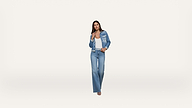 A woman stands facing forward, wearing a blue denim jacket and flared jeans with a white top and sandals, against a plain white background.