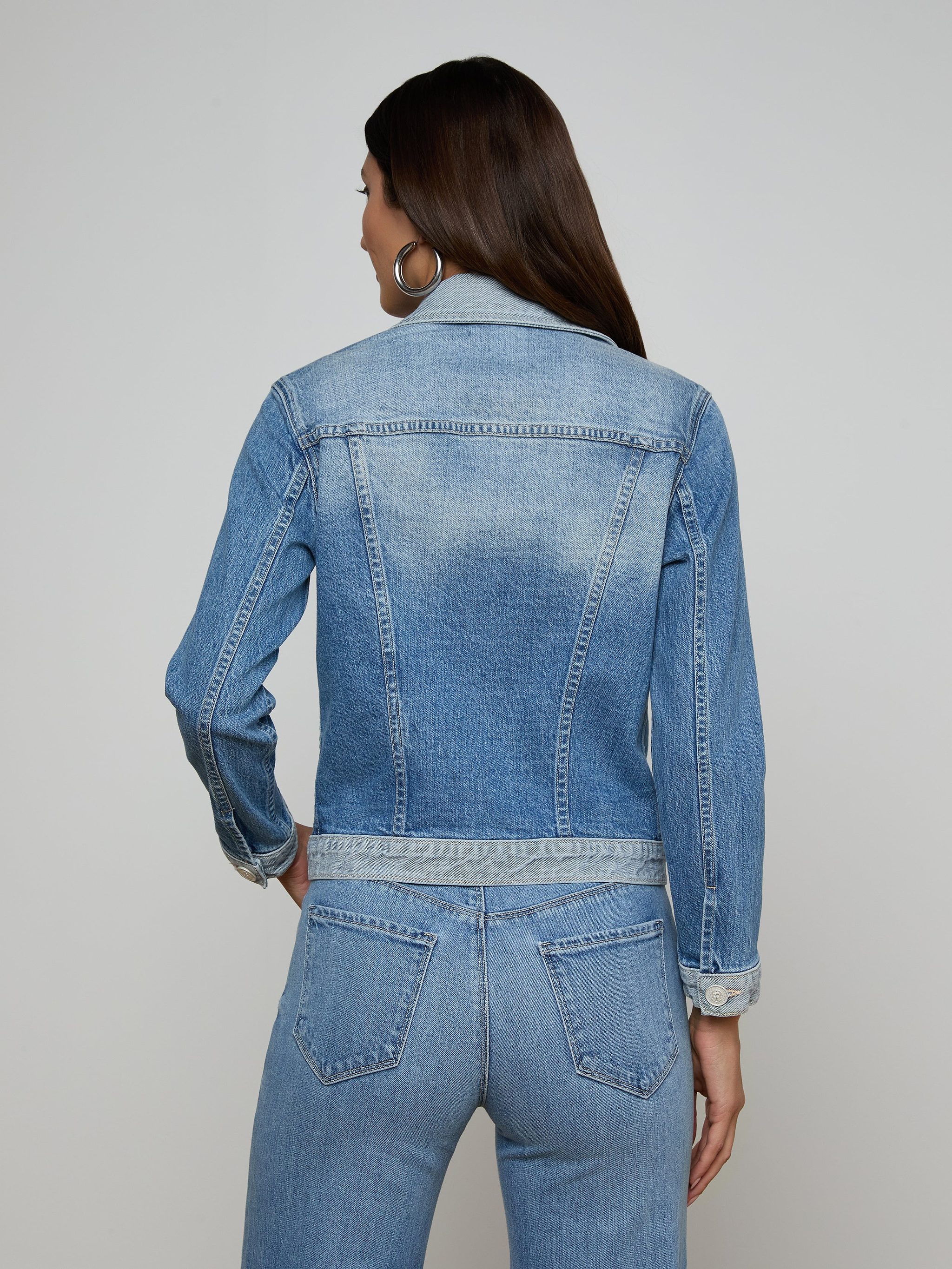 A woman with long dark hair is wearing the Maven Jean Jacket with three-quarter sleeves and matching stretch denim jeans. She is facing away from the camera, highlighting the back of her outfit against a simple light background.