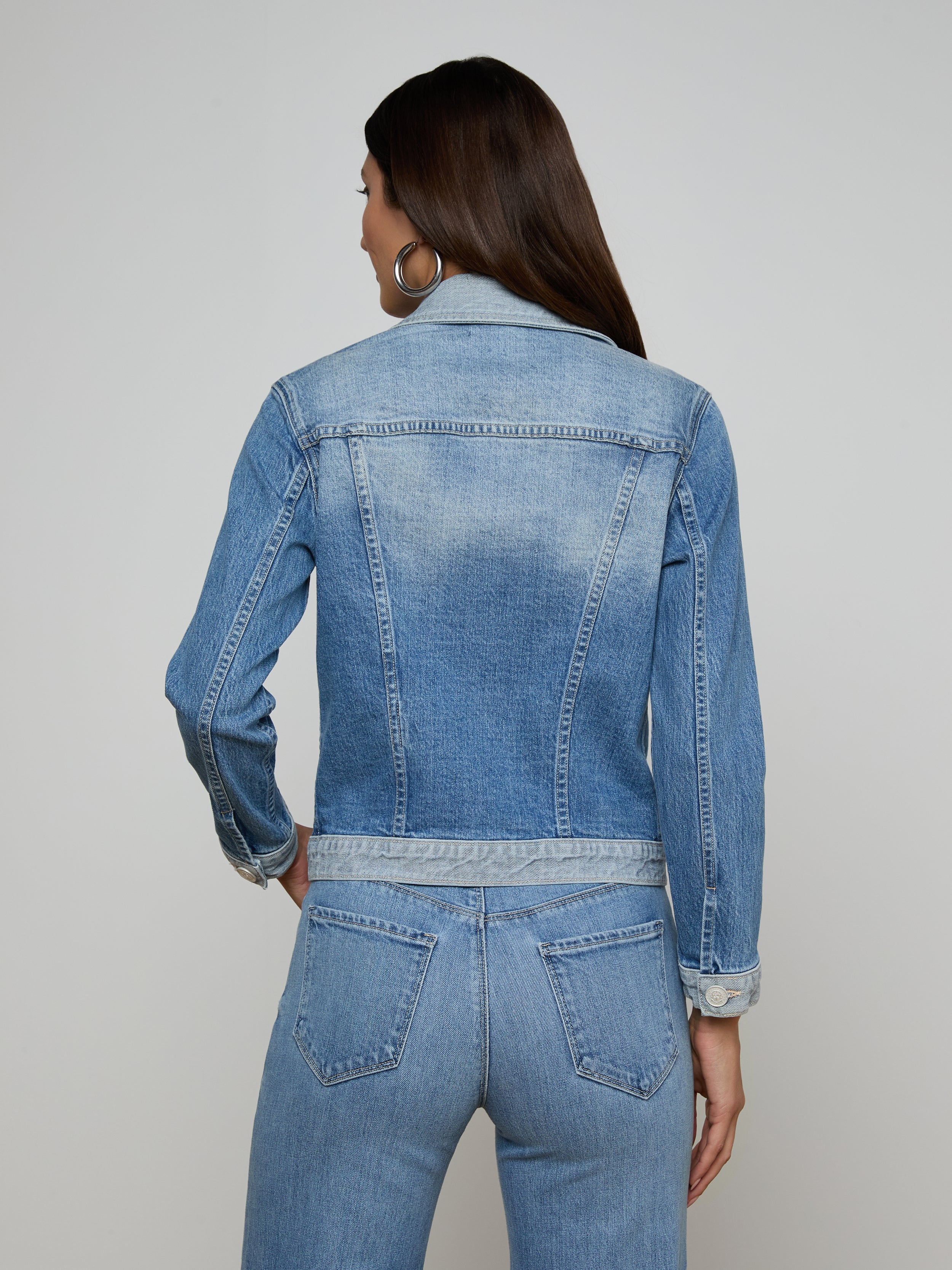 A woman with long dark hair is wearing the Maven Jean Jacket with three-quarter sleeves and matching stretch denim jeans. She is facing away from the camera, highlighting the back of her outfit against a simple light background.