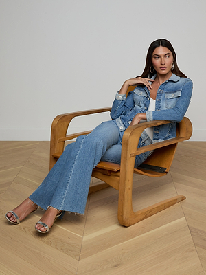 A woman with long brown hair sits in a modern wooden chair, wearing the Maven Jean Jacket and flared light-wash jeans with frayed hems, paired with metallic heeled sandals. She gazes thoughtfully to the side.