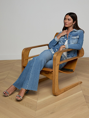 Wearing the Maven Jean Jacket with three-quarter sleeves and light-wash jeans, a woman with long dark hair sits in a wooden chair on a light wood floor, gazing to the side with one hand near her collar and open-toed heels.