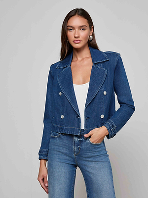 A woman wearing the Maddox Jean Trench Coat over a white top and blue jeans stands against a plain light gray background, looking at the camera with her hand in her pocket.