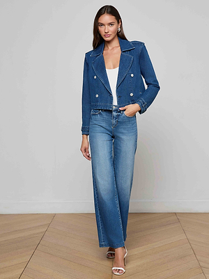 A woman stands indoors on a wooden floor, wearing the Maddox Jean Trench Coat over a white top, blue wide-leg jeans, and white heeled sandals. She looks at the camera with her left hand in her pocket.