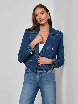 A woman with long brown hair poses against a white wall, wearing the Maddox Jean Trench Coat over a white top and blue jeans, with one hand touching her jacket.